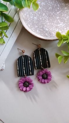 two pairs of black and purple flower earrings on top of a white table next to green plants