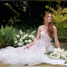 a woman in a wedding dress sitting on the ground with white flowers and greenery behind her