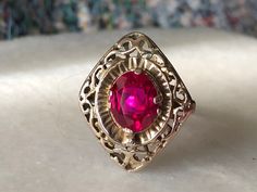 This stunning vintage ring is a beautiful example of antique jewelry craftsmanship, made of 10k gold and featuring a captivating, vibrant red faceted stone at its center. The stone, rich and intense in color, is securely set within an ornate setting that reflects a blend of late Victorian to Art Deco style, characterized by intricate lacy motifs and a delicate filigree pattern. The combination of the deep red stone and the intricate goldwork creates a striking visual contrast, enhancing the over Antique 14k Gold Ruby Ring With Center Stone, Ornate 14k Gold Ruby Ring For Anniversary, Antique 14k Gold Ruby Ring With Birthstone, Antique Red Ruby Birthstone Ring, Heirloom Style Red Birthstone Ring Stamped 14k, Red Vintage Filigree Ring With Gemstone, Antique Hallmarked Ruby Ring, Antique 14k Stamped Ruby Ring, Red Victorian Filigree Ring For Anniversary