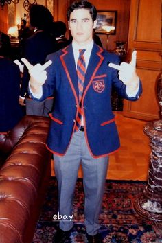 a man in a suit and tie holding two fingers up to the side while standing next to a couch