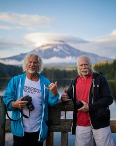 two men standing next to each other on a pier near water with mountains in the background