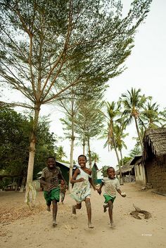 three young children running in the dirt near some trees and shacks with thatched roof