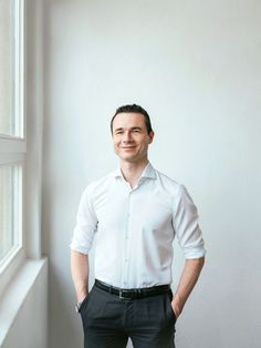 a man standing in front of a window wearing a white shirt and black pants with his hands in his pockets