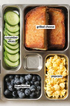 an open lunch box with different foods in it, including bread, cucumber slices, blueberries, and popcorn