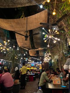 people sitting at tables in a restaurant with lights hanging from the ceiling and plants growing on the walls