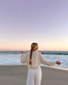 a woman standing on top of a sandy beach next to the ocean with her arms outstretched