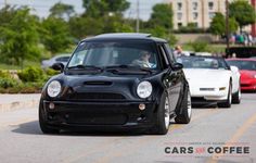 a small black car is driving down the street with other cars behind it and people in the background