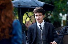 a young man in a suit and tie is talking to another person with an umbrella behind him