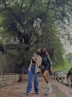 two young women standing next to each other on a brick walkway under a large tree