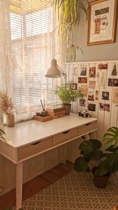 a white desk with plants and pictures on the wall in front of it next to a window