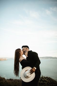 a man and woman are kissing by the ocean with a cowboy hat on their head