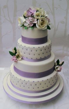 a three tiered wedding cake with purple and white flowers on the top, sitting on a table