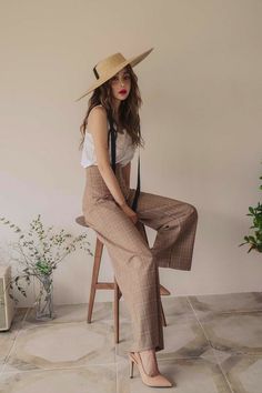 a woman sitting on top of a wooden chair wearing a straw hat and wide legged pants