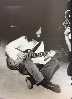 a man sitting on the ground playing a guitar and another person holding an instrument behind him