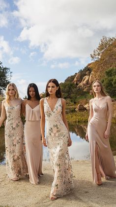 three women in long dresses standing next to each other on the beach with trees and water behind them