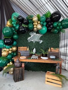 a table topped with lots of green and gold balloons next to a wall covered in greenery