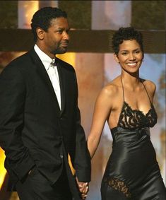 a man and woman in formal wear holding hands at an awards ceremony, one is wearing a black evening gown