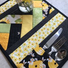 a place mat with yellow and black quilts, silverware and flowers in a vase