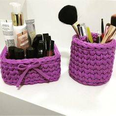 two purple baskets with makeup brushes in them on a white counter top next to each other