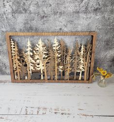 a wooden frame with trees cut out of it on a white wood table next to a vase filled with yellow flowers