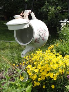 a white tea pot hanging from the side of a planter filled with yellow flowers