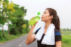 a woman drinking from a green bottle while standing on the side of a road in front of trees