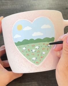 someone is holding a coffee mug with a heart painted on the front and side, while another hand holds a marker