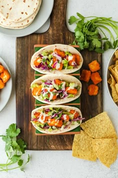 four tacos on a cutting board with tortilla chips and carrots in the background