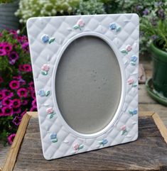 a white frame with pink and blue flowers is sitting on a wooden table next to potted plants