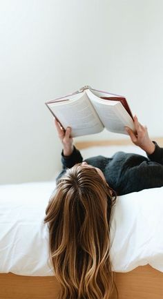 a woman laying on top of a bed holding an open book over her head and reading it