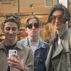 three young men standing next to each other in front of a store window looking at their cell phones