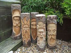 four carved wood faces are sitting on the ground in front of a wooden bench and some plants