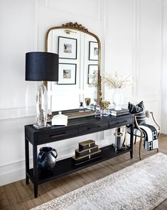 a black console table with a mirror, lamp and vase on it in front of a white wall