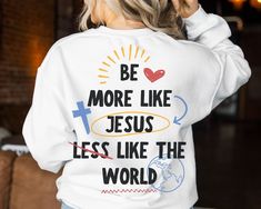 a woman wearing a white sweatshirt that says be more like jesus less like the world