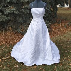 a white wedding dress is on display in front of some trees and grass with fallen leaves