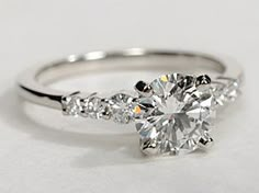 a white gold engagement ring with three stones on the band and a round brilliant cut diamond in the center