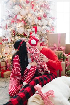 a woman holding a baby in front of a christmas tree with presents on the floor