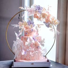 a three tiered cake with pink and purple flowers on top is displayed in front of a window