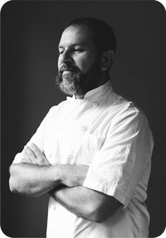 a man with his arms crossed standing in front of a black background wearing a chef's coat