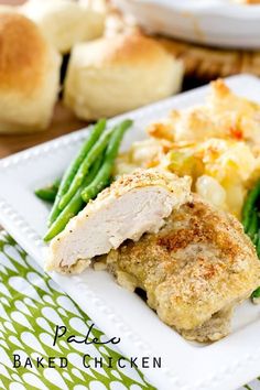 a white plate topped with chicken, green beans and potatoes next to breaded rolls