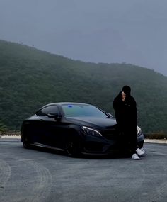 a man standing next to a black car on top of a parking lot with mountains in the background