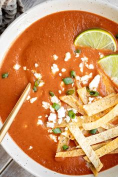 a white bowl filled with red soup and topped with tortilla chips, lime wedges and sour cream