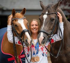 a woman standing next to two horses with their heads on each other's shoulders