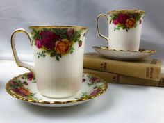 two white cups and saucers with flowers on them sitting next to a stack of books