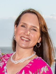 a woman in a pink dress standing next to the ocean and smiling at the camera