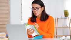 a woman sitting at a table with a laptop and books in front of her, reading a book