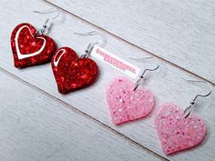 three glitter heart shaped earrings on white wooden surface with name tag attached to the earwires