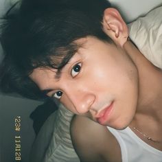 a young man with black hair and white shirt looking at the camera while laying on a bed