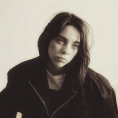 a black and white photo of a woman with long hair wearing a jacket, looking at the camera