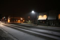 an empty street at night with snow on the ground and buildings lit up by street lights